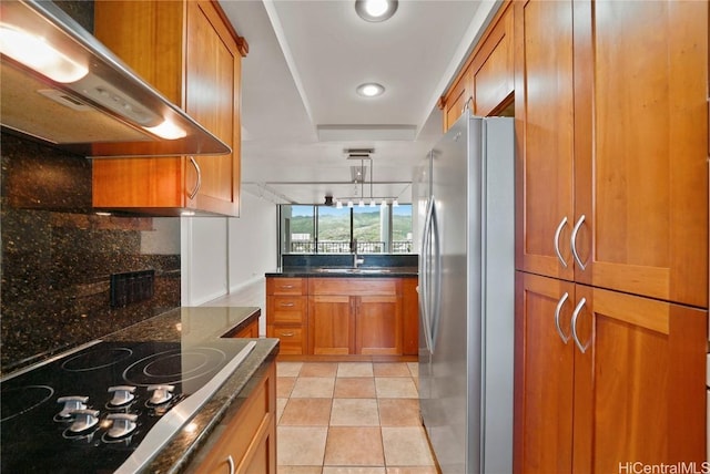 kitchen with brown cabinetry, freestanding refrigerator, a sink, under cabinet range hood, and black electric cooktop