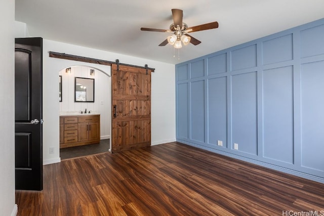 unfurnished bedroom featuring dark hardwood / wood-style floors, connected bathroom, sink, ceiling fan, and a barn door