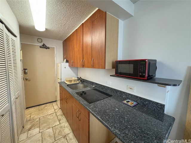 kitchen with a textured ceiling, black electric cooktop, a sink, brown cabinetry, and stone tile flooring