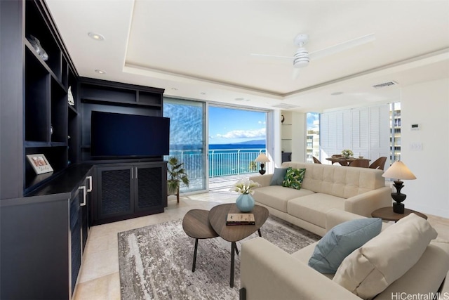 tiled living room featuring built in shelves, a wall of windows, ceiling fan, and a tray ceiling