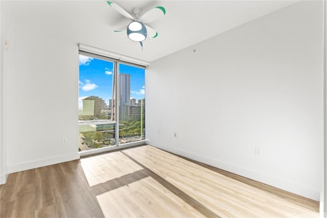 unfurnished room featuring wood finished floors, a ceiling fan, baseboards, a view of city, and a wall of windows