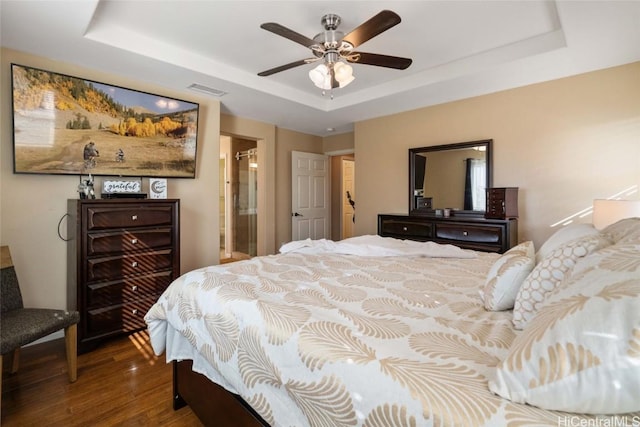 bedroom with a raised ceiling, visible vents, a ceiling fan, ensuite bath, and wood finished floors