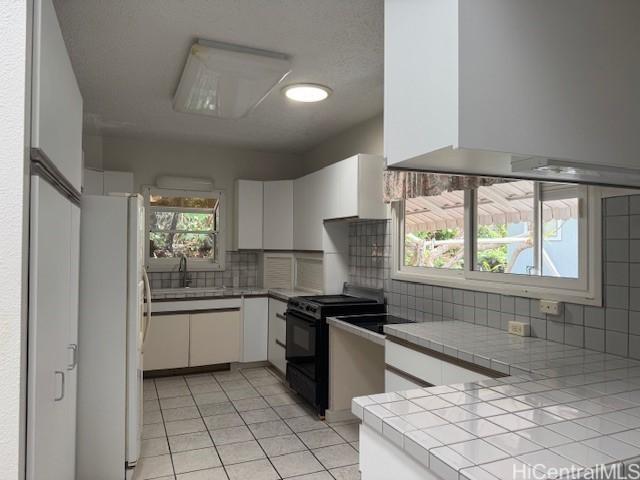 kitchen featuring freestanding refrigerator, white cabinets, black range oven, and tile countertops