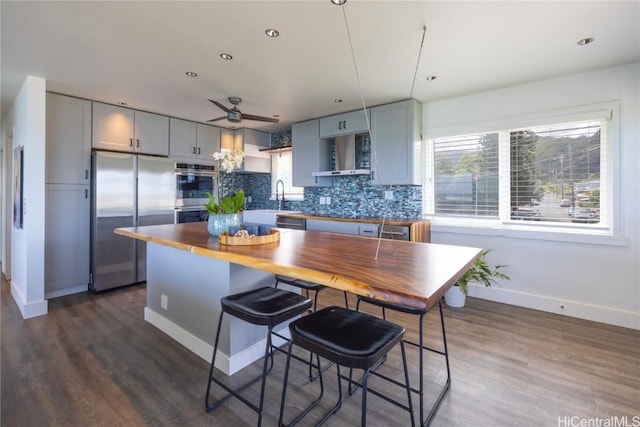 kitchen with dark wood finished floors, stainless steel appliances, backsplash, a sink, and wall chimney range hood