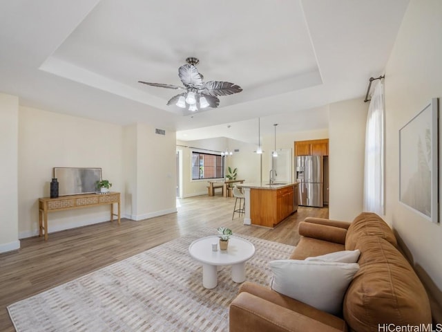 living room featuring ceiling fan, a raised ceiling, sink, and light wood-type flooring