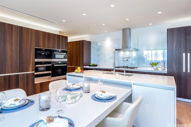 kitchen with an island with sink, wall chimney range hood, modern cabinets, and light countertops