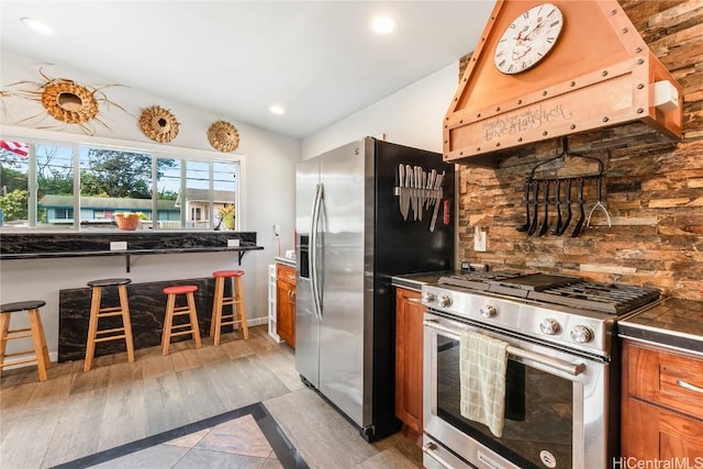 kitchen with dark countertops, brown cabinetry, appliances with stainless steel finishes, and ventilation hood