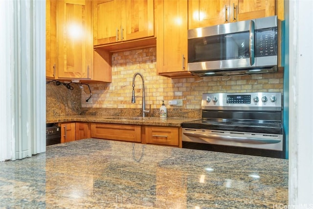 kitchen featuring brown cabinetry, appliances with stainless steel finishes, dark stone countertops, a sink, and backsplash