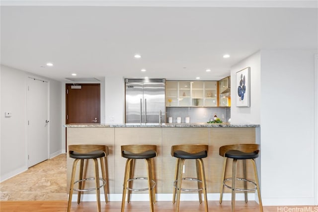 kitchen with light stone counters, glass insert cabinets, stainless steel built in fridge, a peninsula, and a kitchen bar