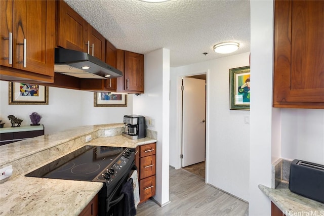 kitchen with a textured ceiling, under cabinet range hood, black electric range, brown cabinets, and light wood finished floors