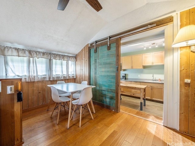 dining space featuring a textured ceiling, a barn door, wood walls, vaulted ceiling, and light wood finished floors