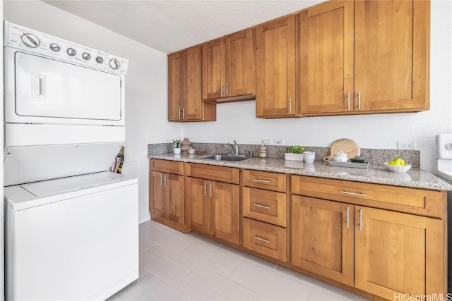 kitchen featuring brown cabinets, light stone countertops, a textured ceiling, stacked washing maching and dryer, and a sink