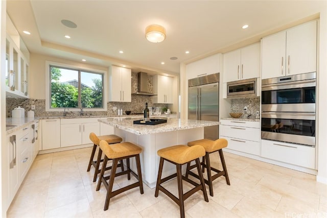 kitchen with a kitchen island, built in appliances, light stone countertops, wall chimney range hood, and a kitchen bar