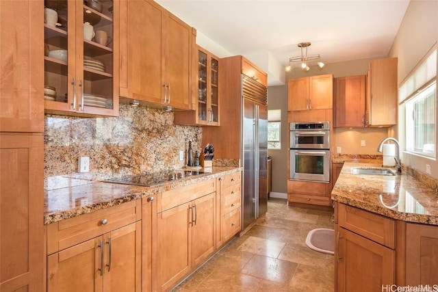 kitchen featuring decorative backsplash, glass insert cabinets, light stone counters, stainless steel appliances, and a sink