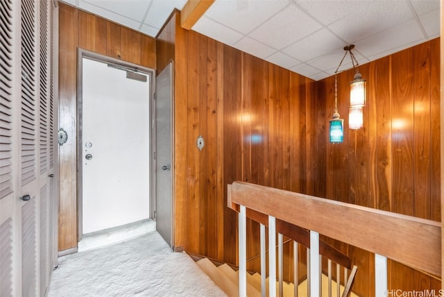 corridor featuring a paneled ceiling, wood walls, carpet flooring, and an upstairs landing