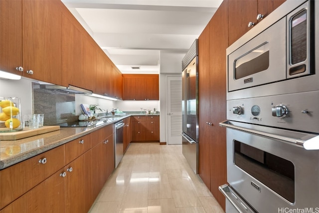 kitchen featuring brown cabinetry, light stone countertops, light tile patterned floors, and built in appliances