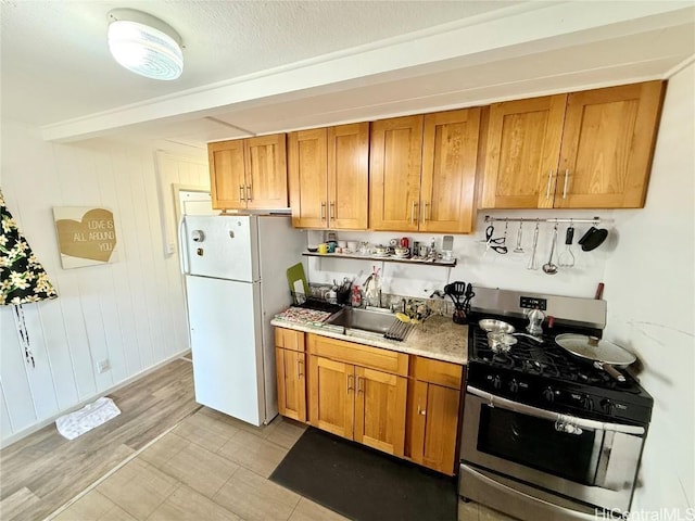 kitchen featuring stainless steel range with gas cooktop, light countertops, brown cabinetry, freestanding refrigerator, and a sink