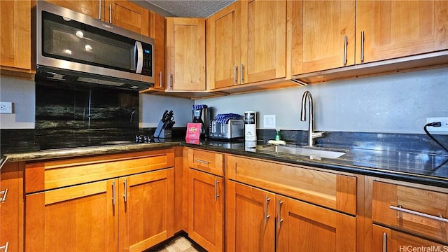 kitchen with stainless steel microwave, brown cabinetry, a sink, and dark stone counters