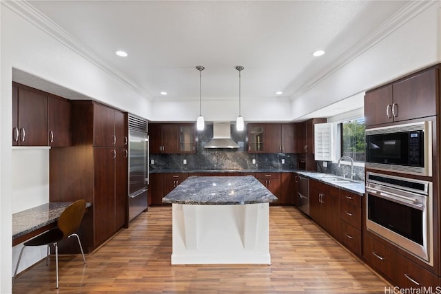 kitchen featuring built in appliances, light wood-style flooring, a sink, wall chimney range hood, and dark stone countertops