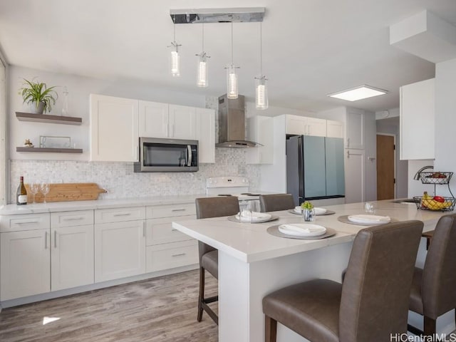 kitchen featuring a breakfast bar area, stainless steel appliances, light countertops, wall chimney range hood, and open shelves