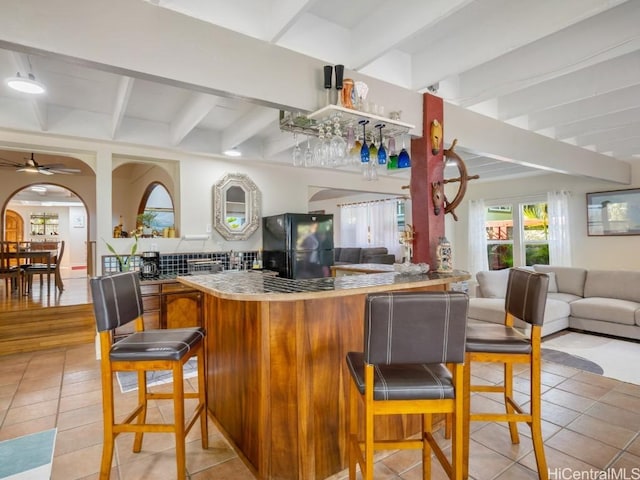 bar with arched walkways, beam ceiling, light tile patterned floors, freestanding refrigerator, and ceiling fan