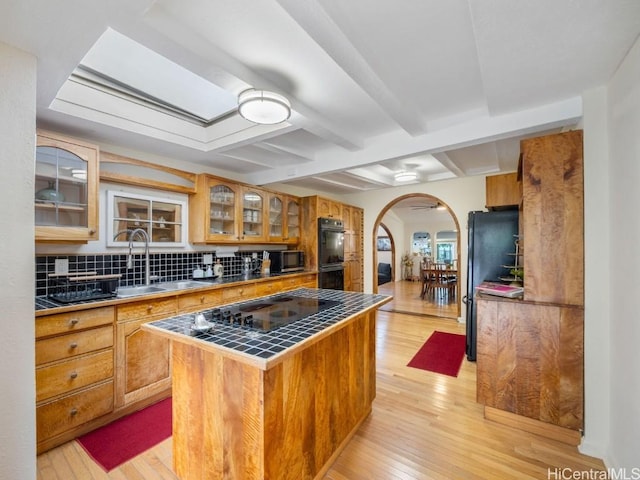 kitchen with light wood finished floors, tile counters, arched walkways, a sink, and beam ceiling