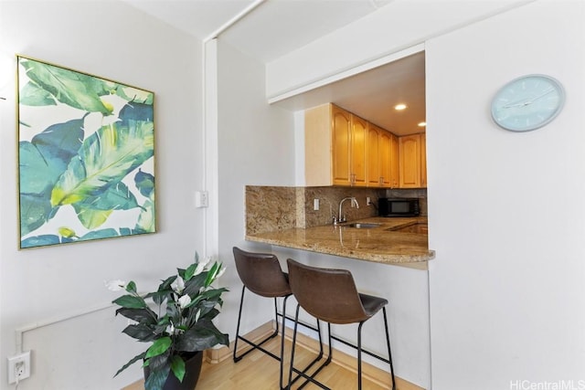 kitchen with light wood finished floors, decorative backsplash, a sink, black microwave, and a kitchen breakfast bar