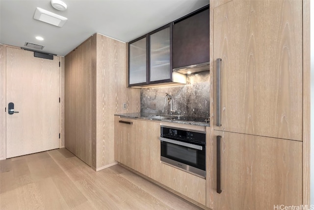 kitchen featuring oven, light wood-style flooring, under cabinet range hood, a sink, and backsplash