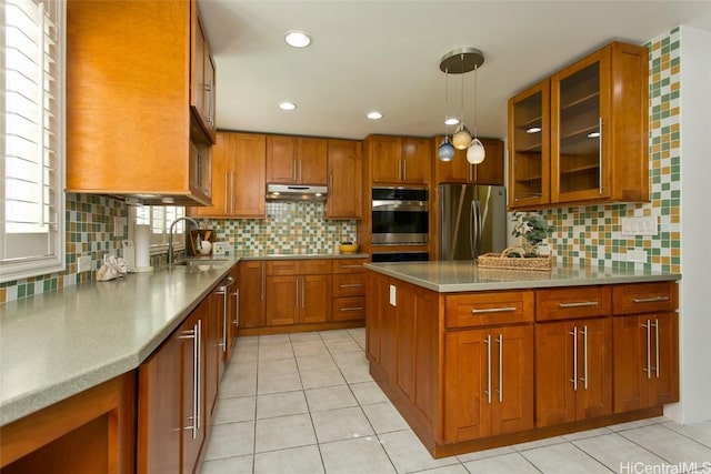 kitchen with under cabinet range hood, stainless steel appliances, a sink, brown cabinetry, and glass insert cabinets