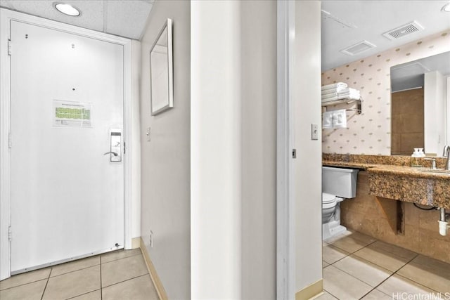 corridor featuring light tile patterned floors, wallpapered walls, baseboards, visible vents, and a sink