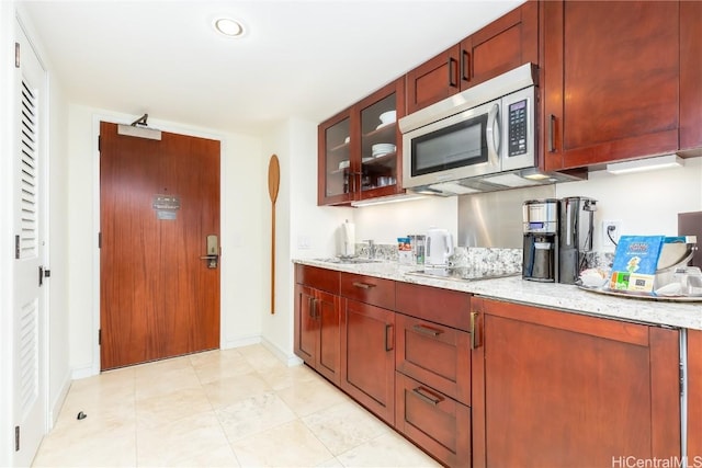 kitchen featuring light stone counters, stainless steel microwave, glass insert cabinets, and baseboards