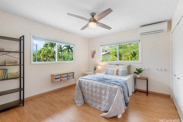 bedroom with multiple windows, a wall mounted air conditioner, light wood-style flooring, and baseboards