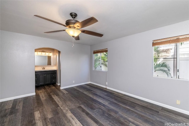 unfurnished bedroom featuring a sink, ensuite bath, arched walkways, baseboards, and dark wood-style flooring