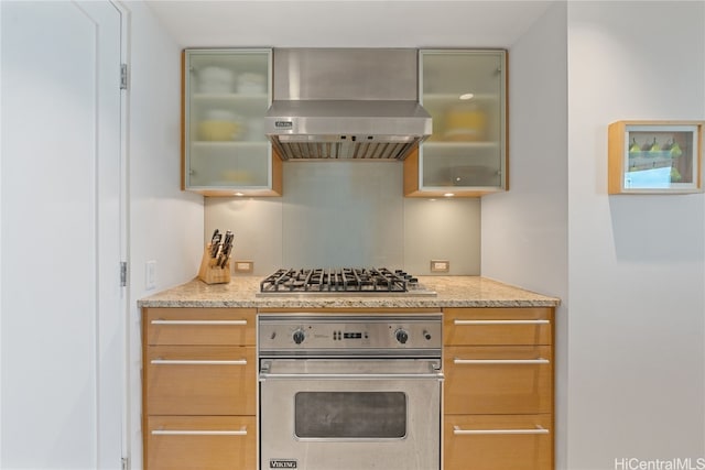 kitchen featuring glass insert cabinets, wall chimney exhaust hood, light stone countertops, and appliances with stainless steel finishes