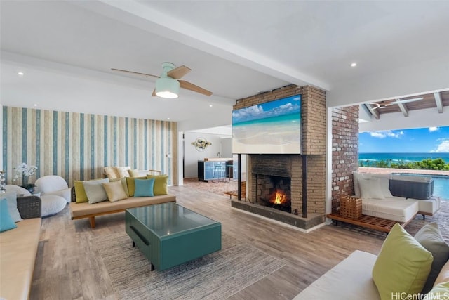 living area featuring beam ceiling, wood finished floors, wallpapered walls, a brick fireplace, and ceiling fan