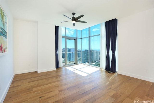 empty room featuring ceiling fan, expansive windows, baseboards, and wood finished floors