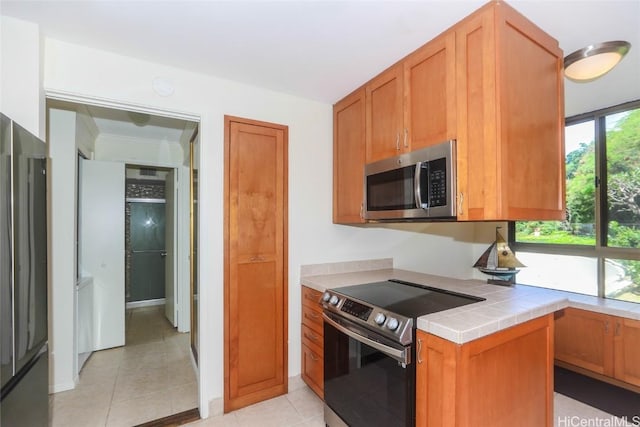 kitchen with tile counters, light tile patterned floors, brown cabinets, and stainless steel appliances