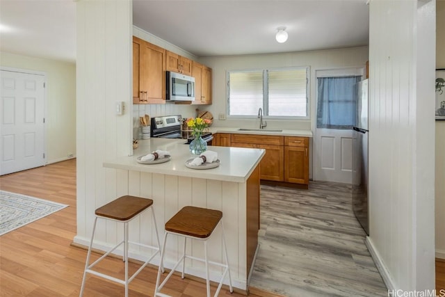 kitchen with a sink, appliances with stainless steel finishes, a peninsula, brown cabinetry, and light countertops