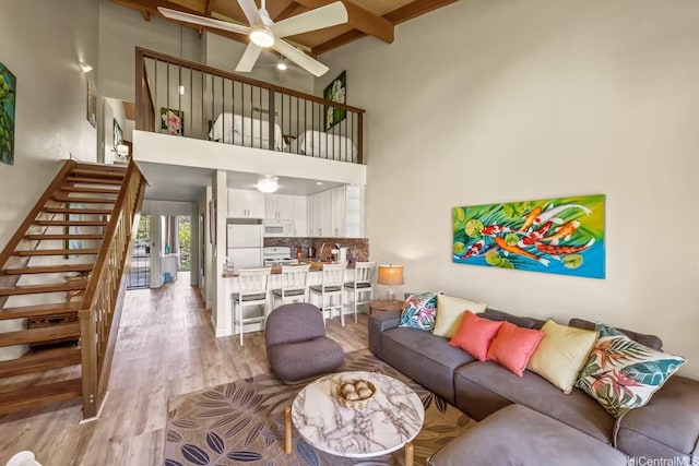 living area featuring a towering ceiling, beamed ceiling, stairway, light wood-type flooring, and a ceiling fan