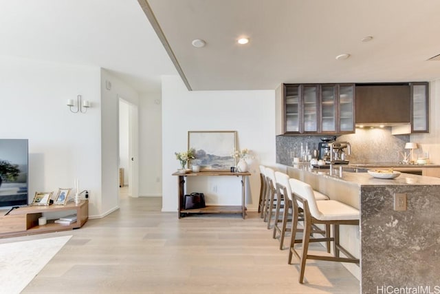 kitchen featuring light wood finished floors, stone counters, decorative backsplash, glass insert cabinets, and a breakfast bar
