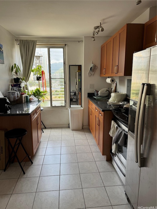 kitchen featuring stainless steel range with gas stovetop, brown cabinets, light tile patterned flooring, dark stone countertops, and white refrigerator with ice dispenser