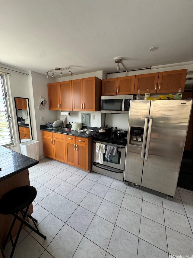kitchen featuring light tile patterned floors, a sink, appliances with stainless steel finishes, dark countertops, and brown cabinetry