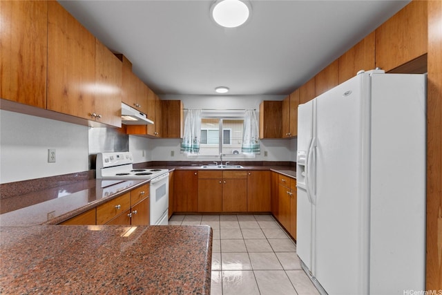 kitchen featuring brown cabinetry, dark countertops, light tile patterned floors, under cabinet range hood, and white appliances