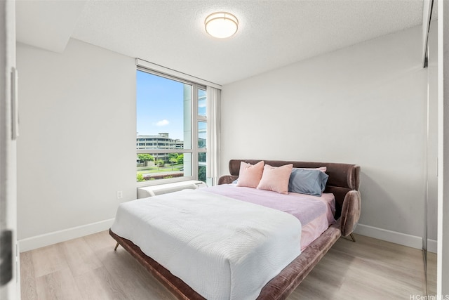 bedroom with a wall of windows, light wood-type flooring, baseboards, a textured ceiling, and a view of city