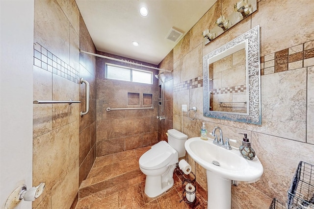 bathroom with a tile shower, a sink, toilet, tile walls, and visible vents