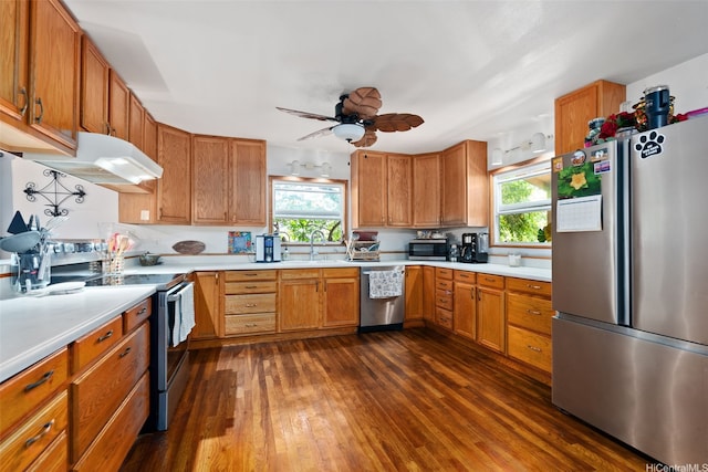 kitchen featuring brown cabinetry, light countertops, under cabinet range hood, stainless steel appliances, and dark wood-type flooring