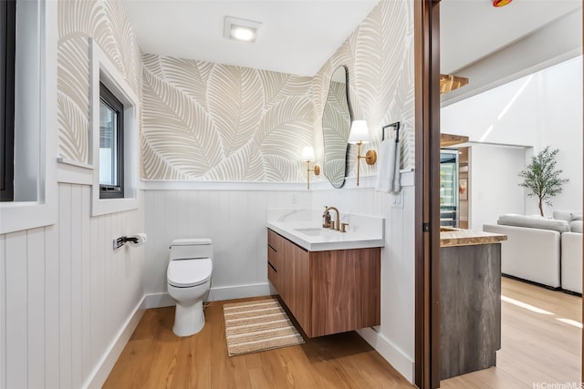 bathroom with toilet, wallpapered walls, wood finished floors, and vanity