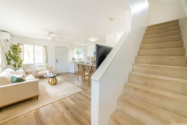 living area with light wood-style flooring, stairs, a wall mounted AC, and ceiling fan