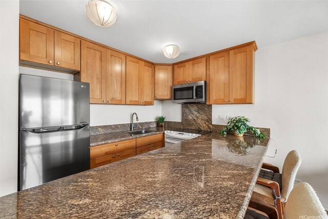 kitchen with appliances with stainless steel finishes, a sink, a peninsula, dark stone countertops, and brown cabinetry