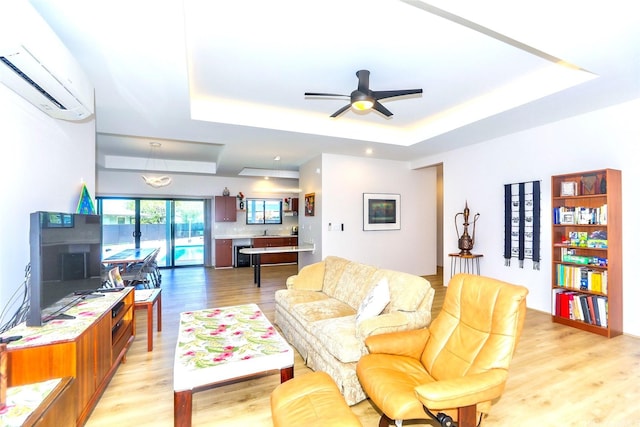 living area with a raised ceiling, light wood-style flooring, a wall mounted AC, and ceiling fan